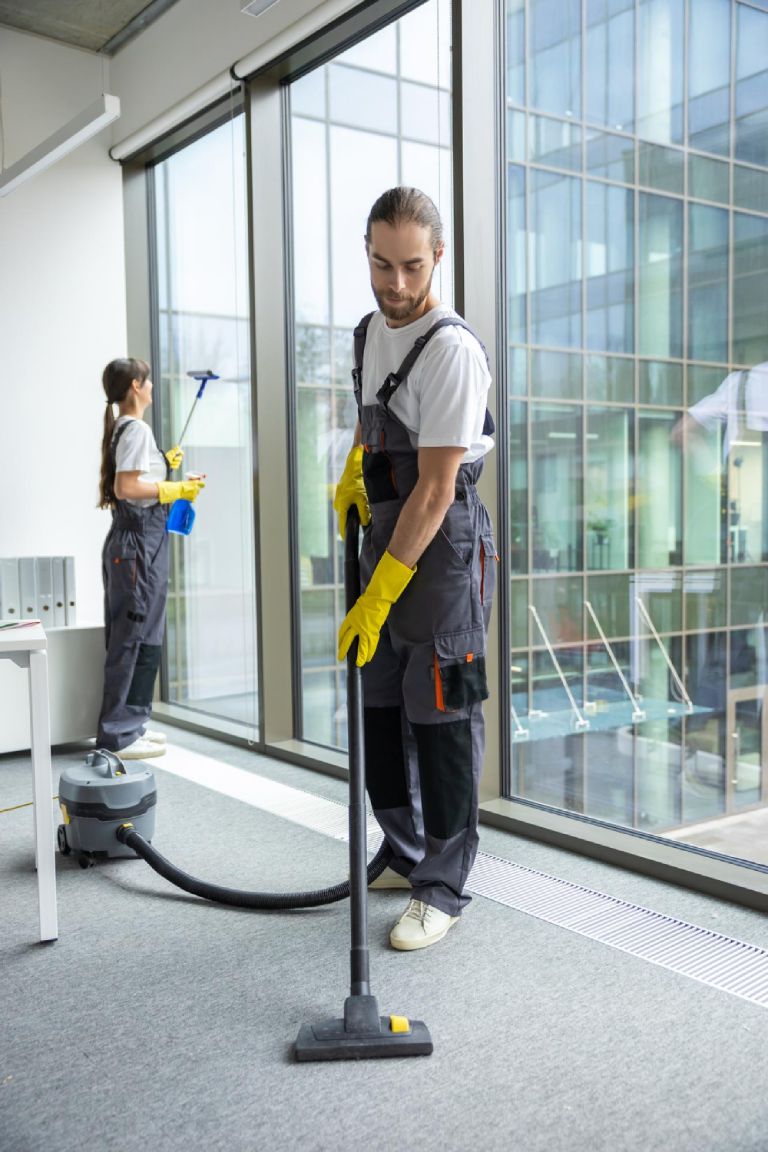 Young man in uniform vacuum cleaning the floor in the conference room urban fresh